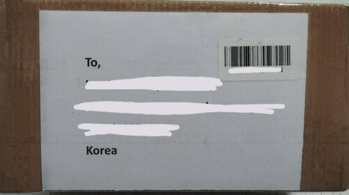 Cardboard box with a white shipping label showing 'To, Korea' and a barcode sticker; recipient details are obscured.