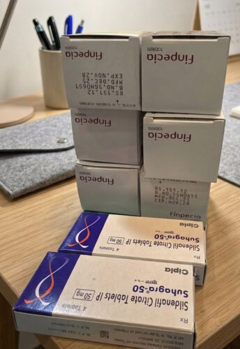 Stack of six white Finpecia boxes stacked in a 2-by-3 arrangement on a wooden desk; two blue Sildenafil citrate boxes lie flat in front.
